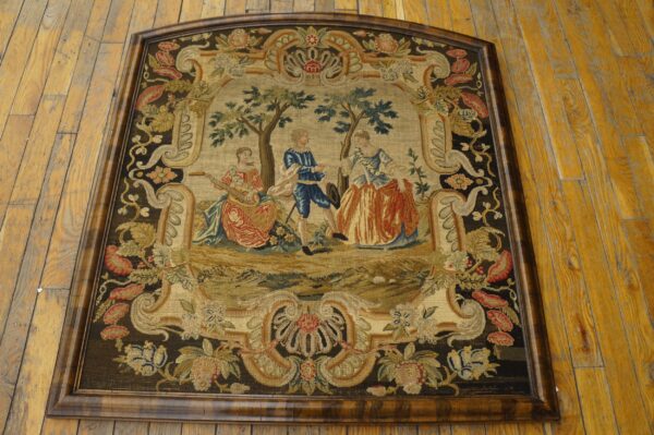 A framed needlepoint tapestry rug featuring a central pastoral scene bordered by dark floral motifs on wood flooring.