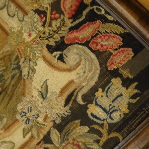 Ornate floral needlepoint textile in gold, red, and beige on a black background framed by dark carved wood.