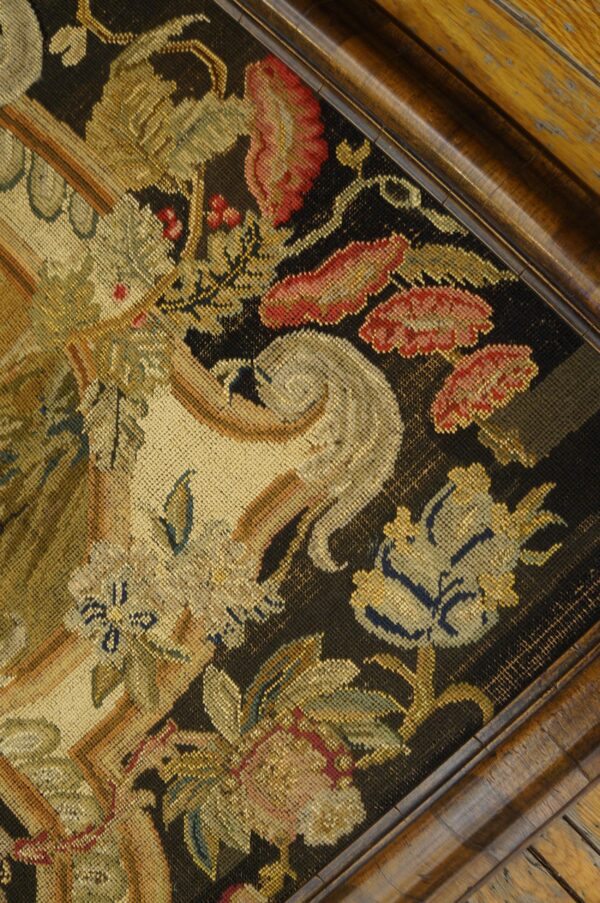 Ornate floral needlepoint textile in gold, red, and beige on a black background framed by dark carved wood.
