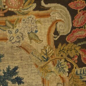 Ornate floral and botanical needlepoint textile in muted reds, greens, and gold against a dark background, set in a wood frame.