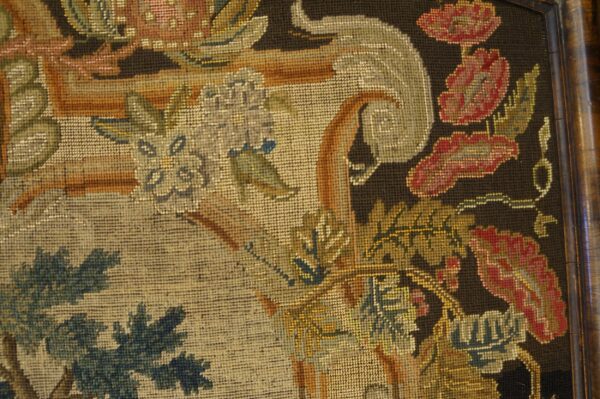 Ornate floral and botanical needlepoint textile in muted reds, greens, and gold against a dark background, set in a wood frame.