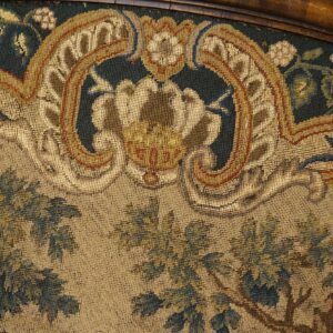 Close-up of a woven textile showing dark green foliage, beige ground, and gold baroque scrollwork under wood trim.