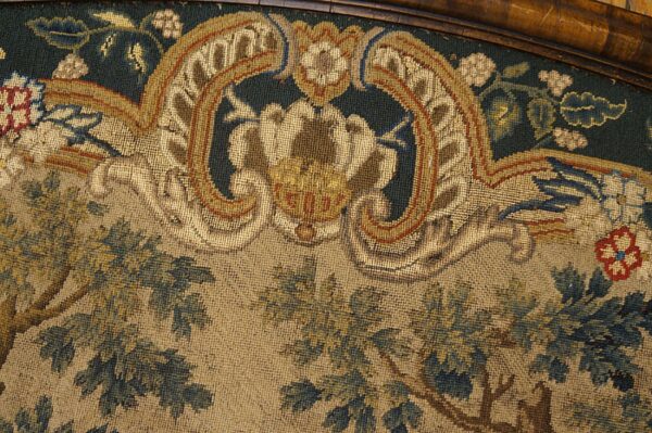 Close-up of a woven textile showing dark green foliage, beige ground, and gold baroque scrollwork under wood trim.