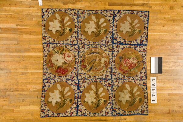 Flatweave rug with a nine-square floral medallion design in tan, blue, and cream on a wood floor.