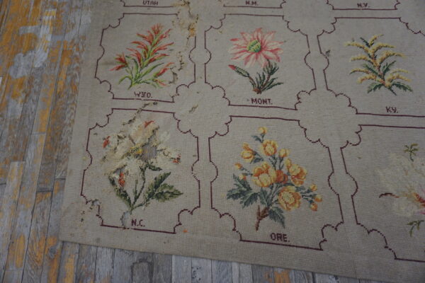 Worn beige low-pile rug with floral and lettered panels rests on heavily peeling gray wood floor.