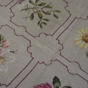 A beige needlepoint rug displaying colorful floral patterns within repeating, scalloped maroon frames.