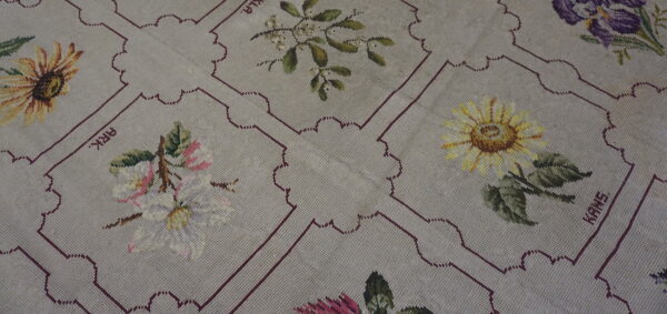 A beige needlepoint rug displaying colorful floral patterns within repeating, scalloped maroon frames.