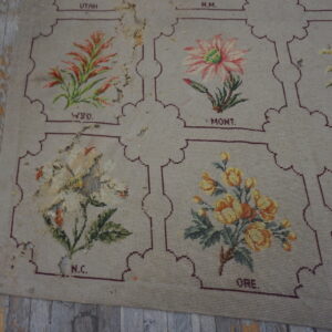 A worn beige needlepoint rug featuring floral panels bordered in maroon, lying on distressed gray and yellow wood floors.