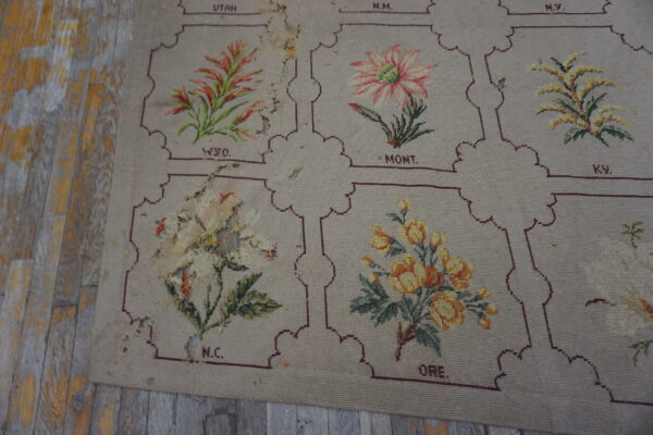 A worn beige needlepoint rug featuring floral panels bordered in maroon, lying on distressed gray and yellow wood floors.