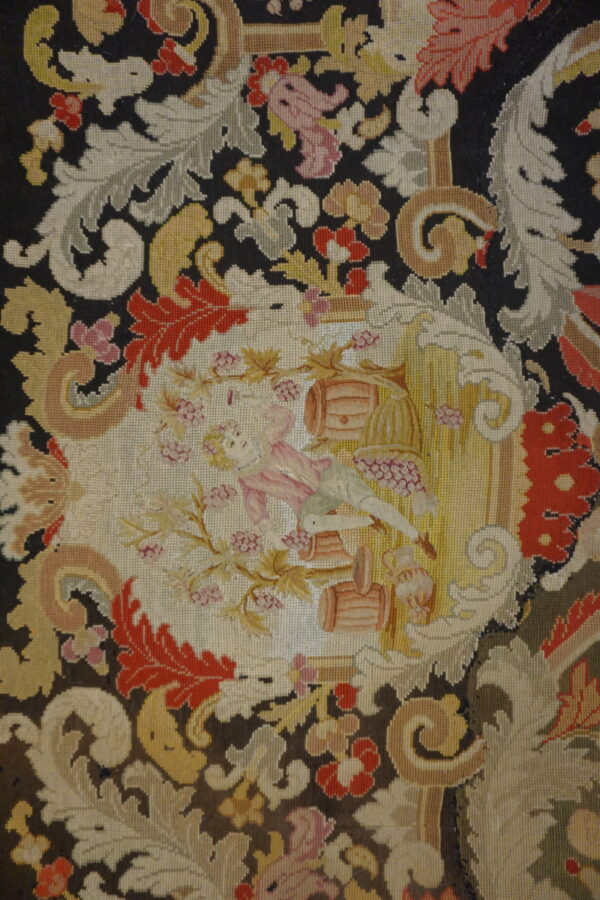 Low-pile needlepoint rug featuring dark ground, colorful baroque foliage, and a central cartouche scene.