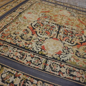 Low-pile ornate rug with black, cream, red, and green classical motifs on wide plank wood flooring.
