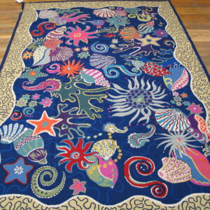 A large, low-pile blue rug features a vibrant, colorful undersea motif and a tan squiggly border on a wood floor.