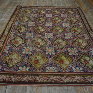 A large low-pile rug with blue, brown, and green diamond trellis pattern featuring floral bouquets rests on worn wood.