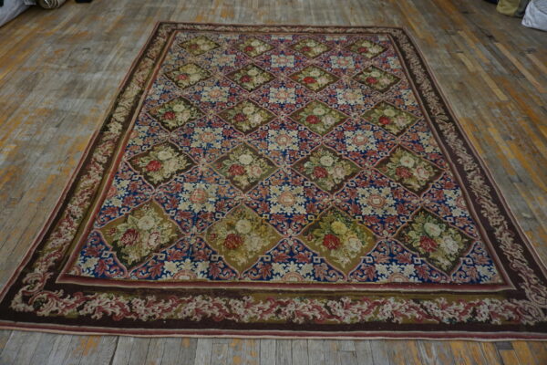A large low-pile rug with blue, brown, and green diamond trellis pattern featuring floral bouquets rests on worn wood.