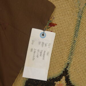 Close-up of a tan textured rug with colorful patterns, partially covered by a brown fabric and white identification tag.