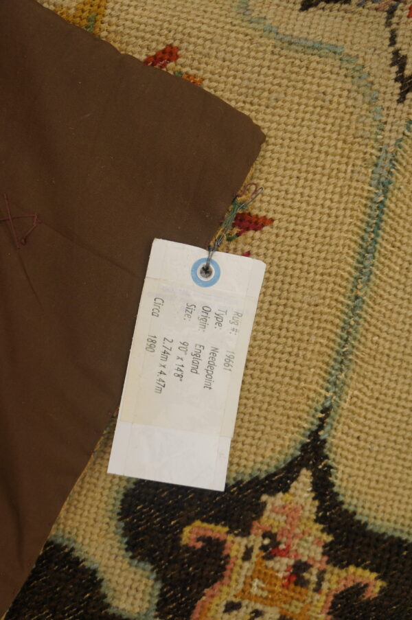 Close-up of a tan textured rug with colorful patterns, partially covered by a brown fabric and white identification tag.