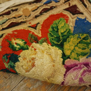 Textured floral hooked rug in red, green, blue, and yellow partially rolled on rustic wood planks.