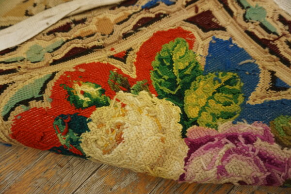 Textured floral hooked rug in red, green, blue, and yellow partially rolled on rustic wood planks.