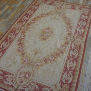 Low-pile rug featuring a large pink floral wreath and red border on weathered hardwood flooring.