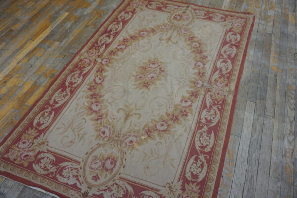 Low-pile rug featuring a large pink floral wreath and red border on weathered hardwood flooring.