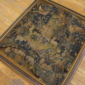 Framed, fine-stitch textile displaying blue, green, and tan figures and trees rests on worn wood flooring.