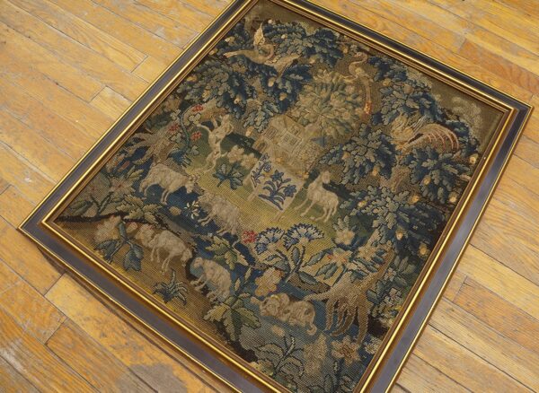 Framed, fine-stitch textile displaying blue, green, and tan figures and trees rests on worn wood flooring.