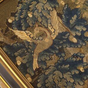 Close-up of a low-pile framed textile featuring a tan bird among dense blue and olive foliage.