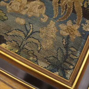 Detail of a framed needlepoint textile showing blue, beige, and green foliage patterns on distressed wood flooring.