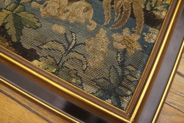 Detail of a framed needlepoint textile showing blue, beige, and green foliage patterns on distressed wood flooring.