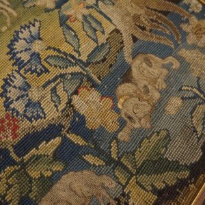 Muted green, blue, and tan needlepoint textile featuring complex floral and animal designs within a gold frame.