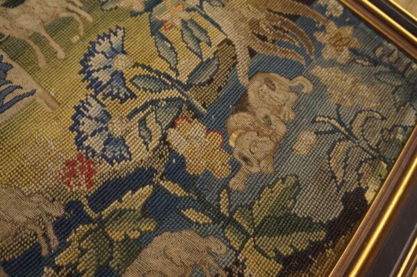 Muted green, blue, and tan needlepoint textile featuring complex floral and animal designs within a gold frame.