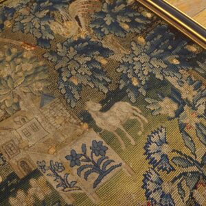 Close-up of a framed flat woven textile featuring blue and gold foliage, animals, and a building on angled wood flooring.