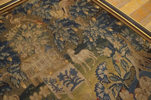 Close-up of a framed flat woven textile featuring blue and gold foliage, animals, and a building on angled wood flooring.