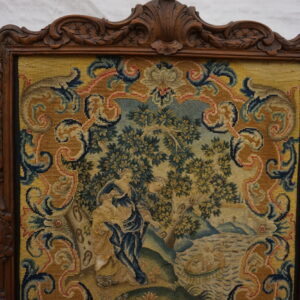 Ornate carved wood frame around a detailed needlepoint panel depicting figures, trees, and gold scrolling borders.