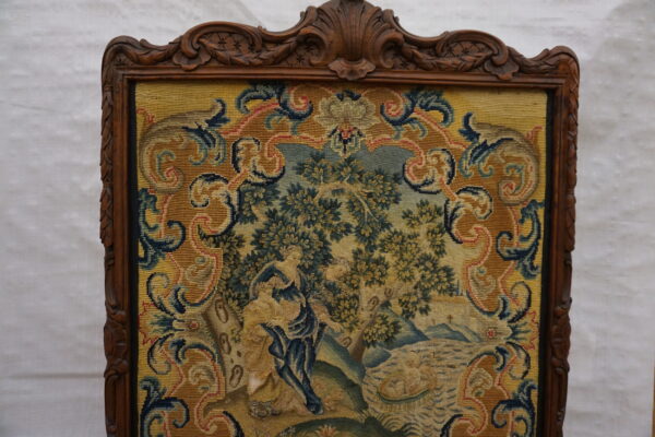 Ornate carved wood frame around a detailed needlepoint panel depicting figures, trees, and gold scrolling borders.