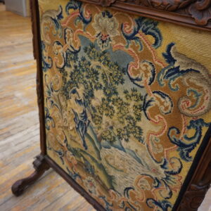 Low-pile tapestry panel with a yellow, blue, and green pastoral scene in a dark carved frame on wood floor.
