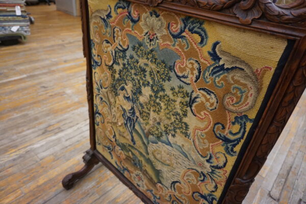 Low-pile tapestry panel with a yellow, blue, and green pastoral scene in a dark carved frame on wood floor.