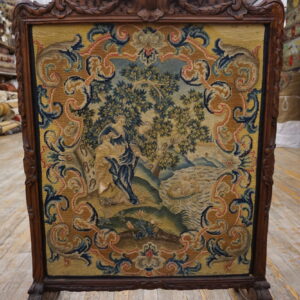 A framed tapestry featuring a blue, green, and gold figural scene, displayed on worn wooden flooring.