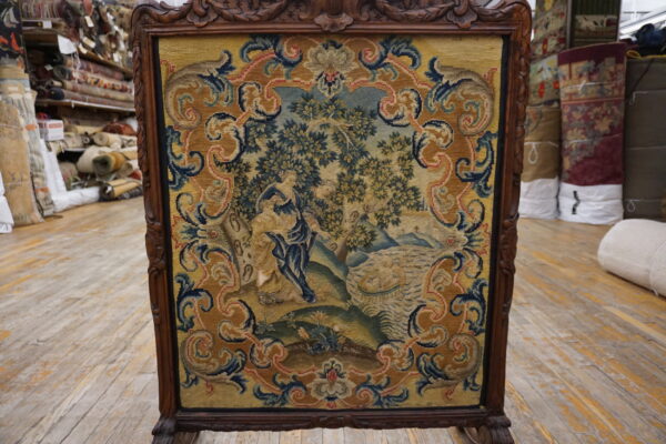 A framed tapestry featuring a blue, green, and gold figural scene, displayed on worn wooden flooring.