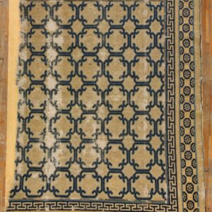 Heavily worn tan rug with dark blue geometric trellis and medallion borders resting on a wooden floor.