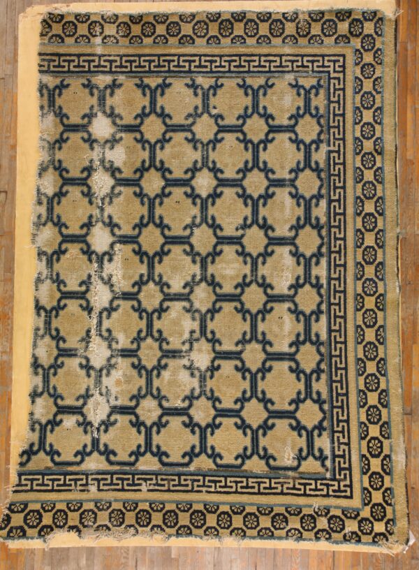 Heavily worn tan rug with dark blue geometric trellis and medallion borders resting on a wooden floor.