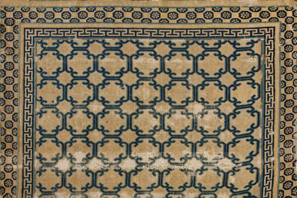 Tan rug with a distressed low pile features a repeating dark blue geometric lattice and greek key border.