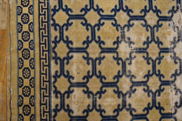 Worn tan rug features dark blue geometric trellis and patterned borders on wood flooring.