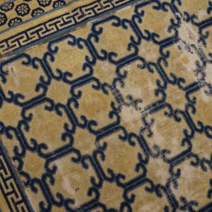 Distressed yellow rug featuring blue geometric lattice and key borders, showing damage on wood floor.