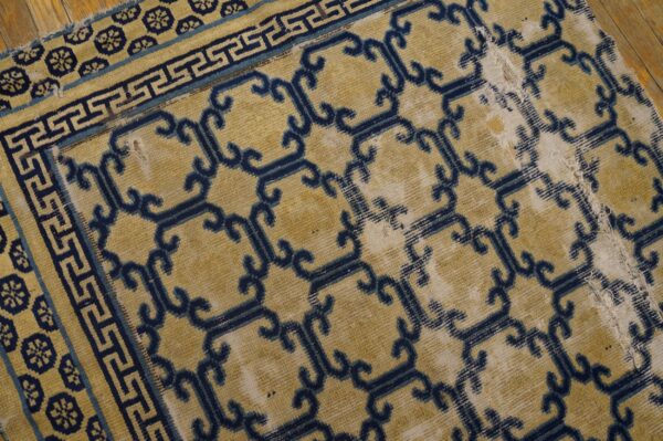 Distressed yellow rug featuring blue geometric lattice and key borders, showing damage on wood floor.