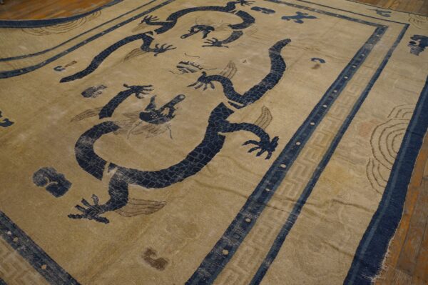 Worn, khaki and navy rug with large dragon motifs and a striped geometric border over wood flooring.