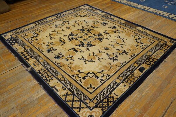 Ornate square rug in cream, gold, and navy blue with geometric borders on wood floor.