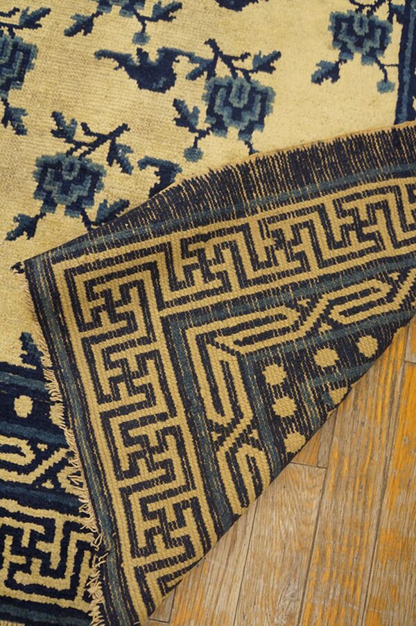 Close-up of a yellow and indigo floral rug folded to show a geometric border on a wooden floor.