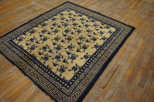 A square pale yellow rug featuring repeating blue florals and geometric borders rests on rustic wood planks.
