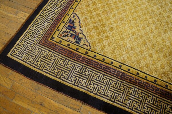 Golden rug featuring a geometric maze border and corner spandrel resting on worn plank wood flooring.